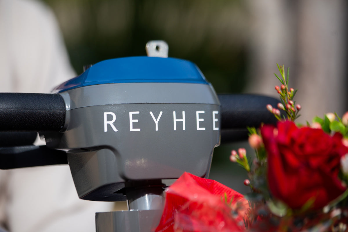 Up close of the Reyhee logo in white on the blue Reyhee Cruiser mobility scooter with red flowers in the front basket