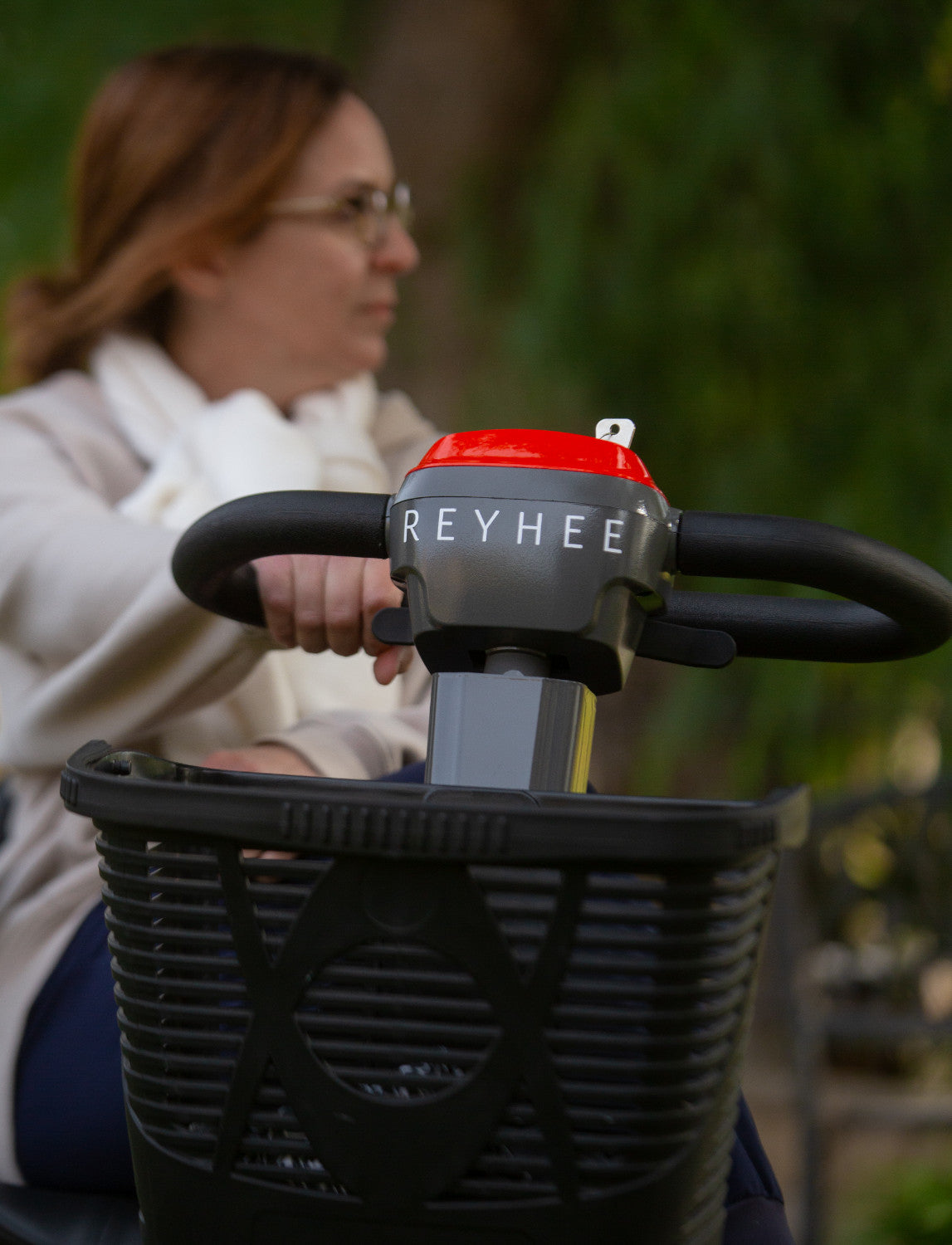 Close up of the front of the red Reyhee Cruiser mobility scooter with a woman's hand on the handle as she looks to the side in the background